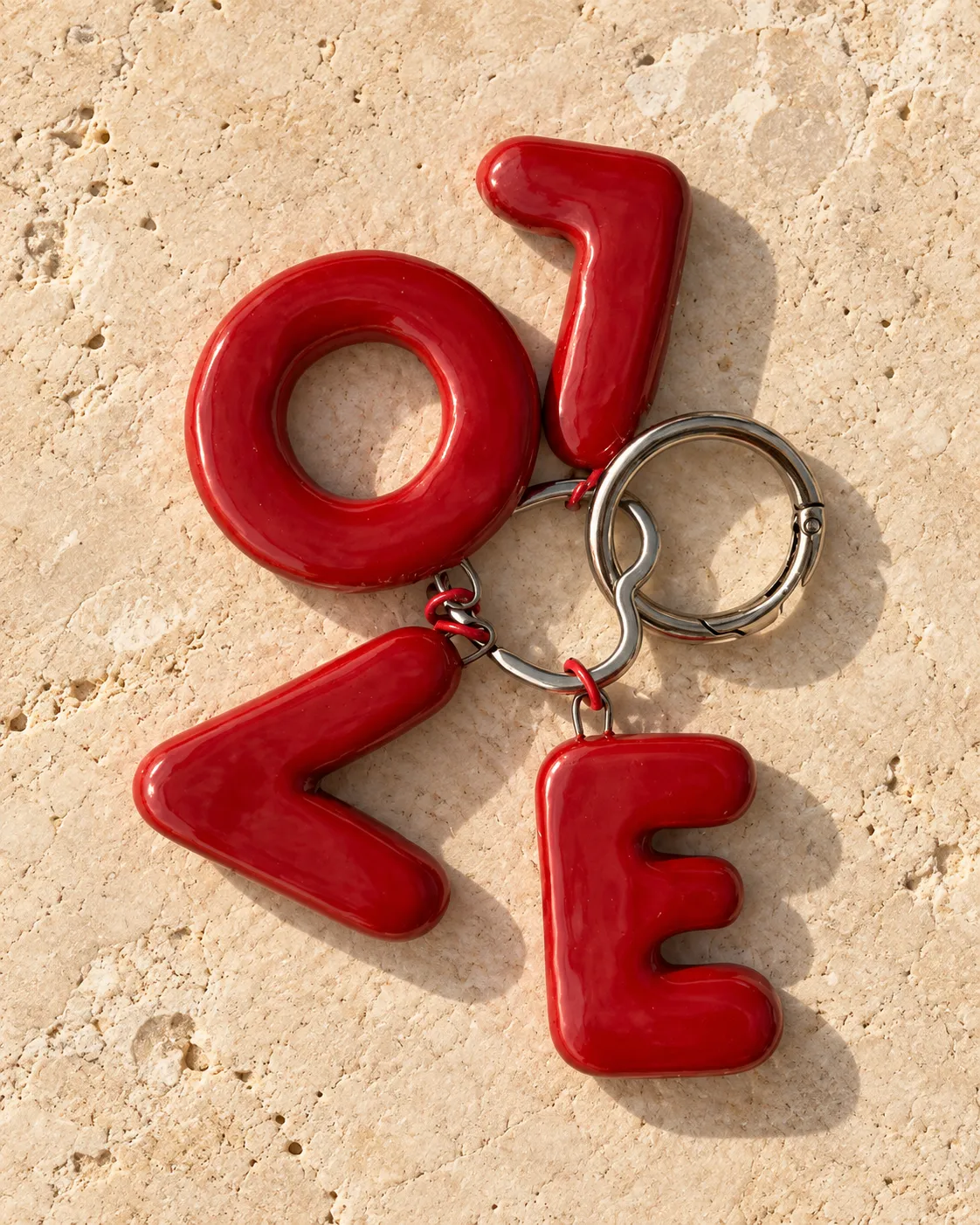 Four glossy red ceramic letters spelling LOVE, clipped to a silver ring, on travertine