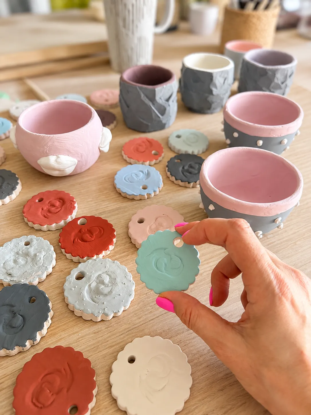 A hand with pink nails holding a turquoise scalloped ceramic medallion, with more medallions in terracotta, blue, grey and white and small pink and grey painted bowls behind