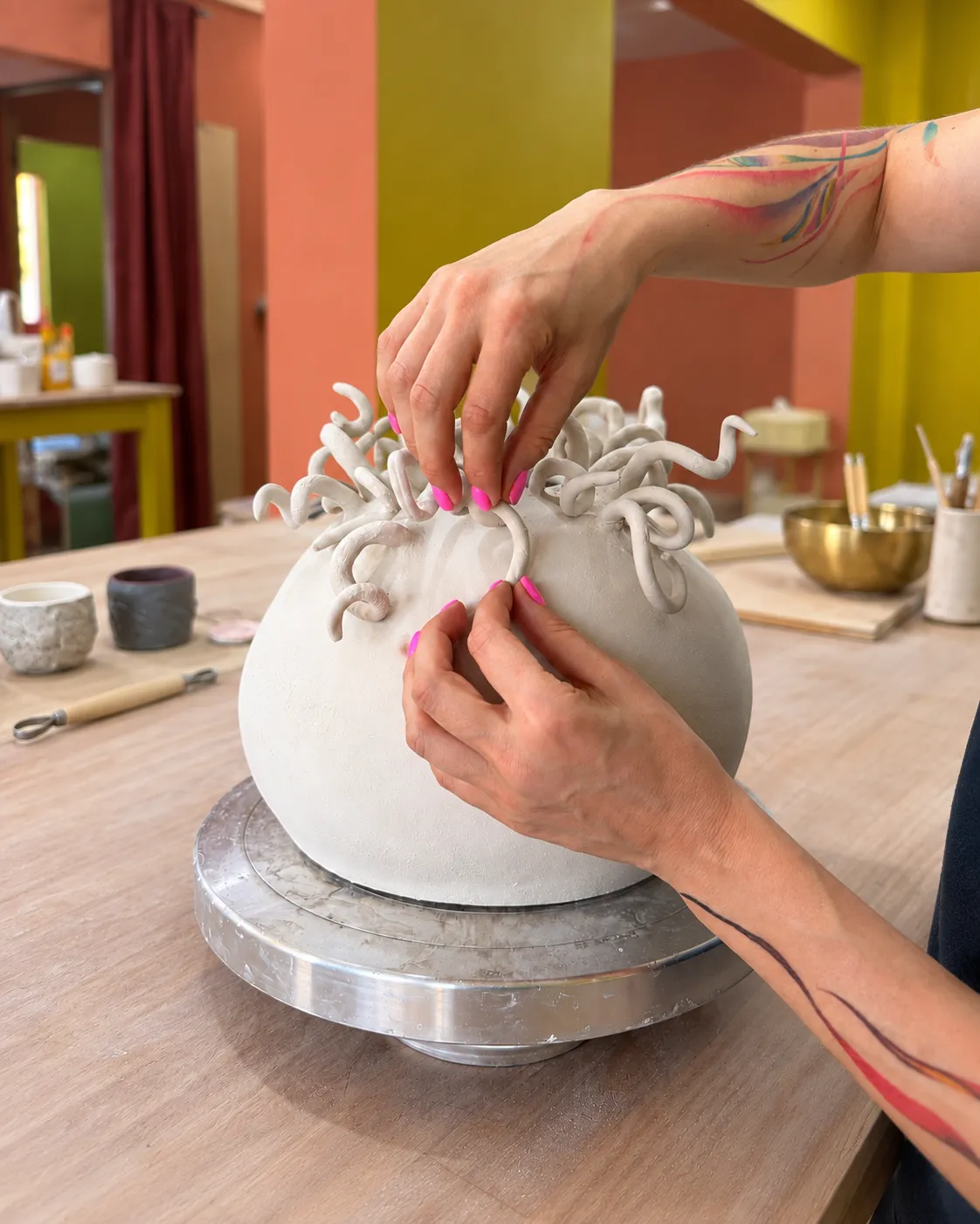 Tattooed hands attaching curled clay tendrils to the top of a large round white vessel on a banding wheel, with tools, a brass bowl and terracotta and mustard walls soft-focused behind