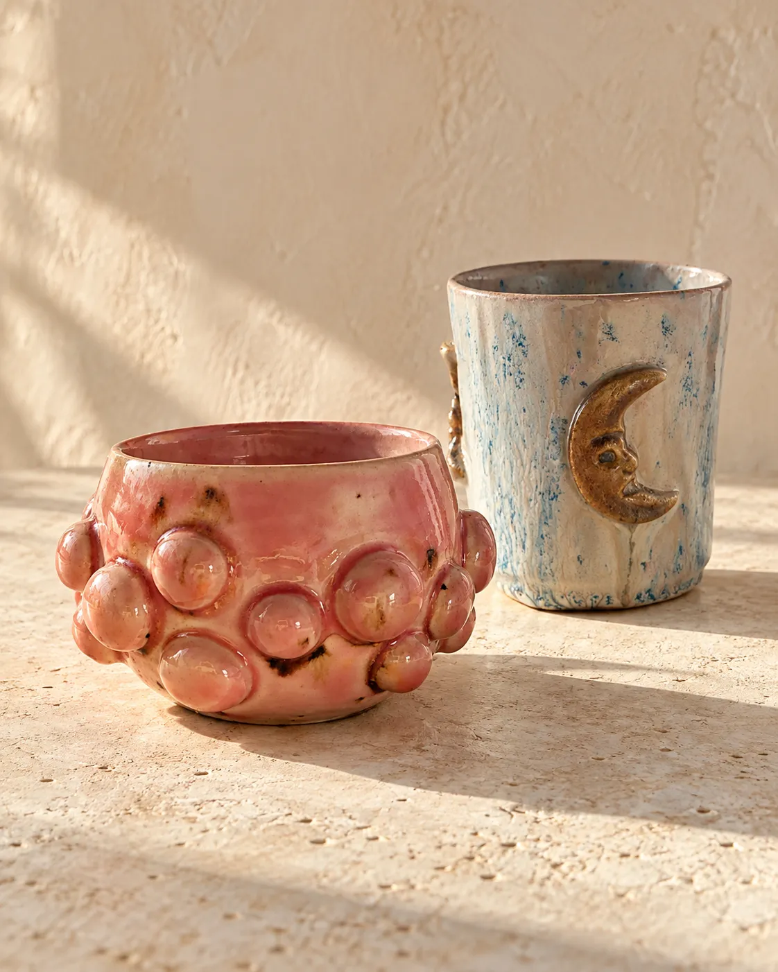 A glazed mug with a raised moon face, standing beside a pink bubble-textured bowl