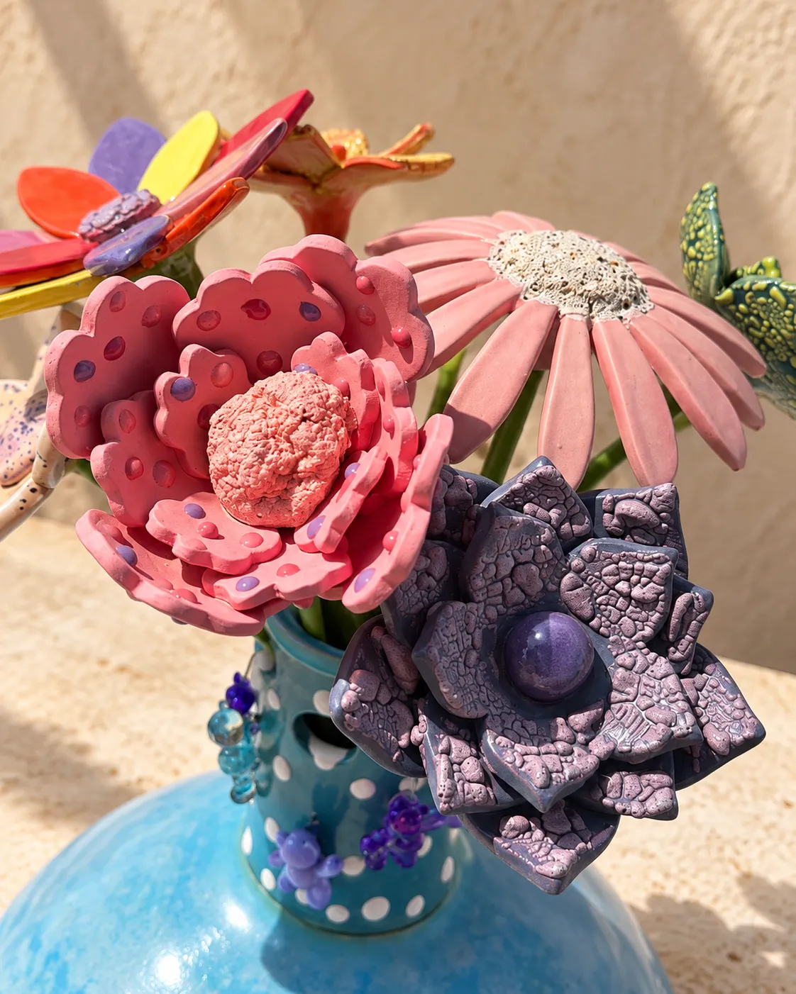 Hand-built ceramic flowers in a turquoise polka-dot vase — a dotted pink peony, a pink daisy with a textured cream centre and a purple crackle-glazed bloom