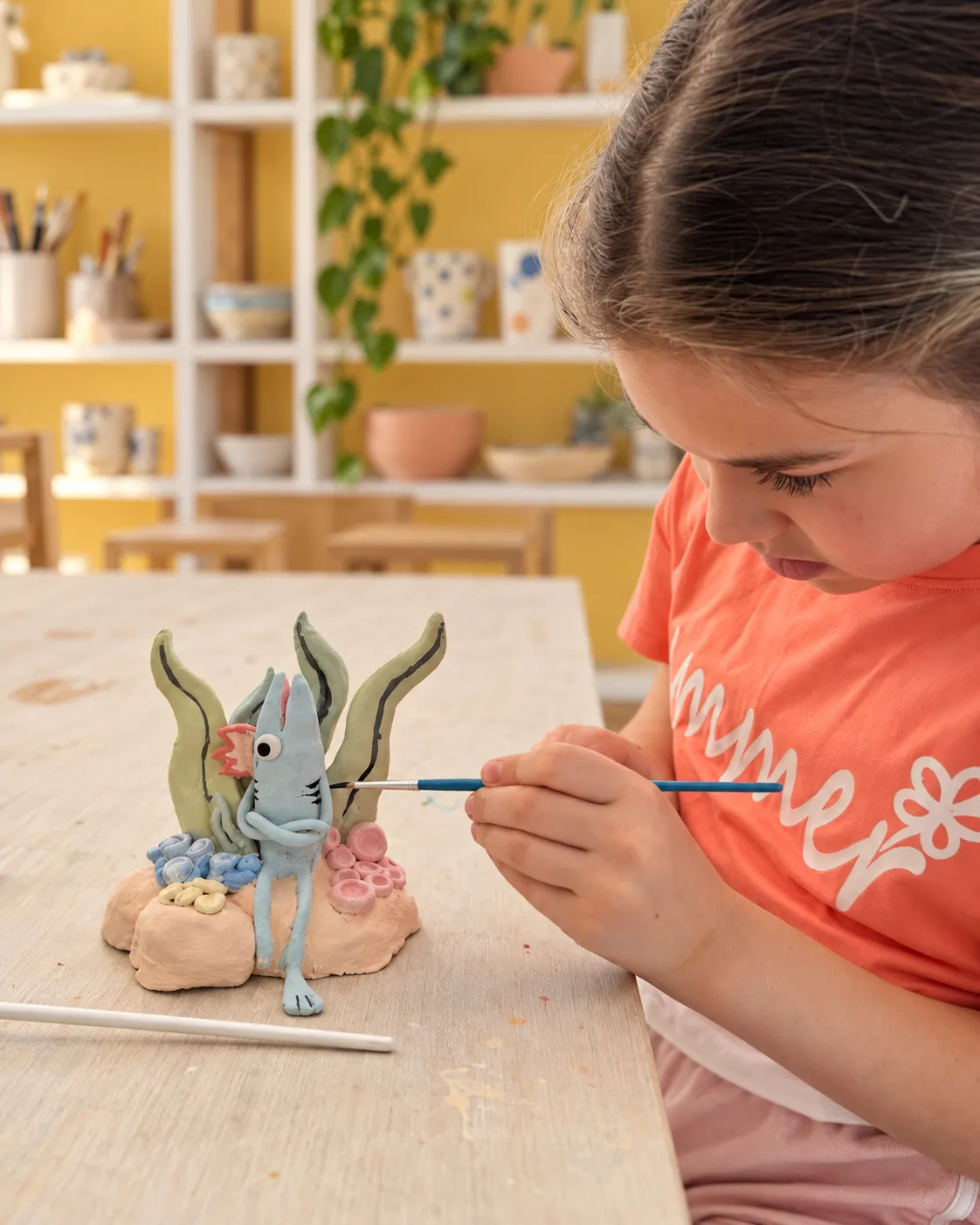 A girl in a coral T-shirt painting a small blue ceramic sea creature among clay seaweed and corals, with studio shelves behind