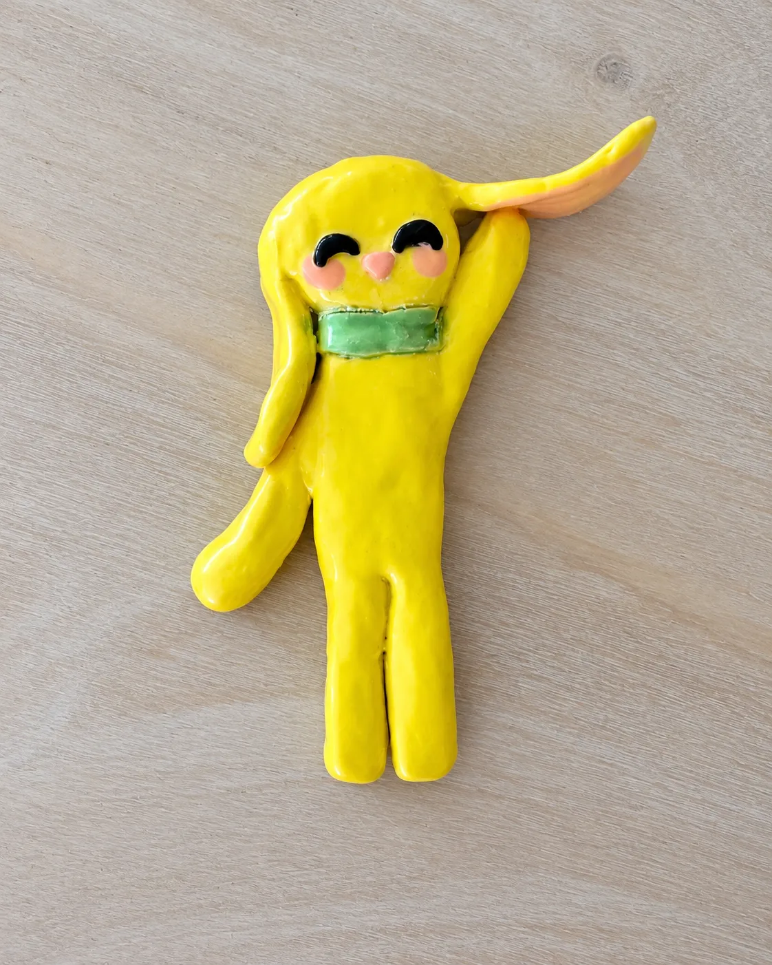A glossy yellow ceramic bunny with a green scarf, closed smiling eyes and rosy cheeks, holding up one long ear, on light wood