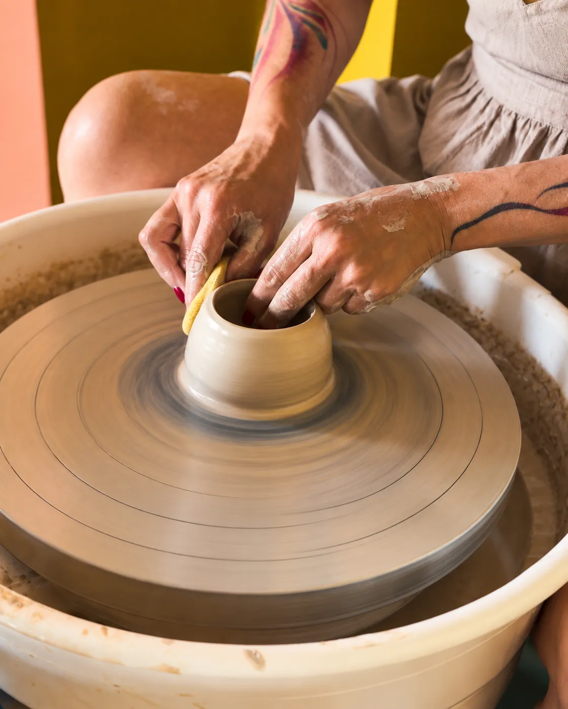Close-up of tattooed, clay-dusted hands shaping a small pot on a spinning wheel with a yellow sponge, a grey linen apron and a terracotta and mustard wall behind