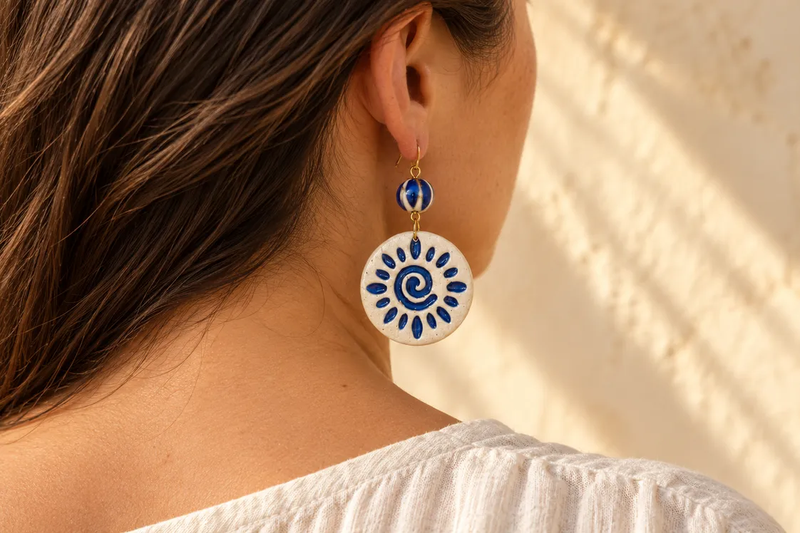 A round white ceramic earring painted with a cobalt-blue spiral sun, hung below a striped blue bead and worn, seen from behind