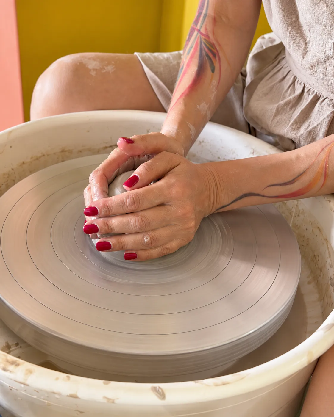 Hands with red nails centring a ball of clay on a spinning pottery wheel, colourful line tattoos on both forearms and a yellow wall behind